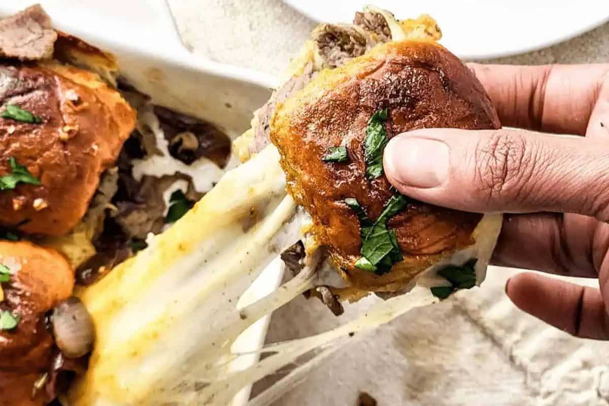 A hand pulls a steak and cheese slider with melted cheese from a dish, perfect for crowd-pleasing recipes. The golden bun is garnished with herbs.