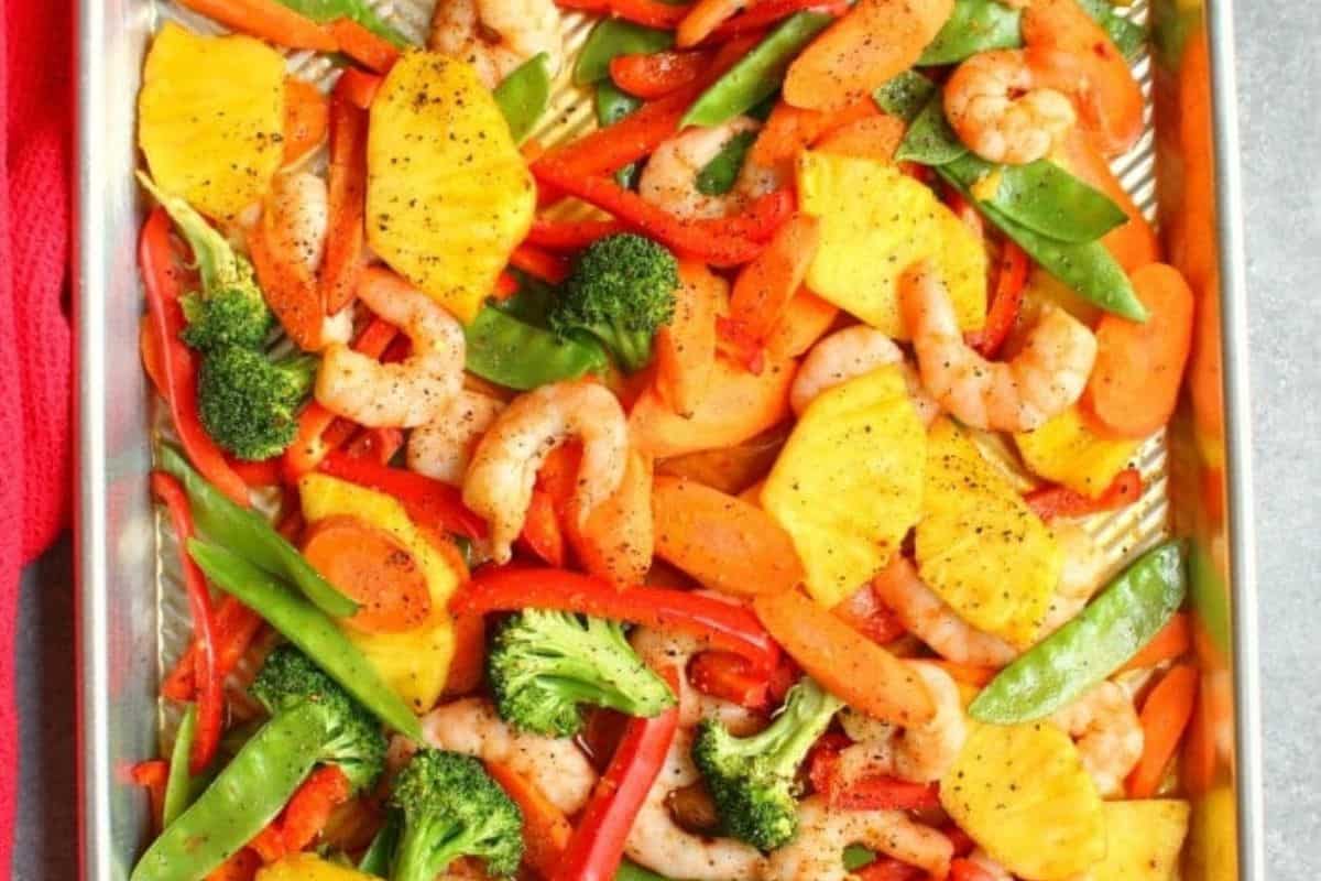Colorful sheet pan with shrimp, sliced pineapples, bell peppers, carrots, broccoli, and snap peas seasoned with pepper for a restaurant-quality dish that's easy to cook.