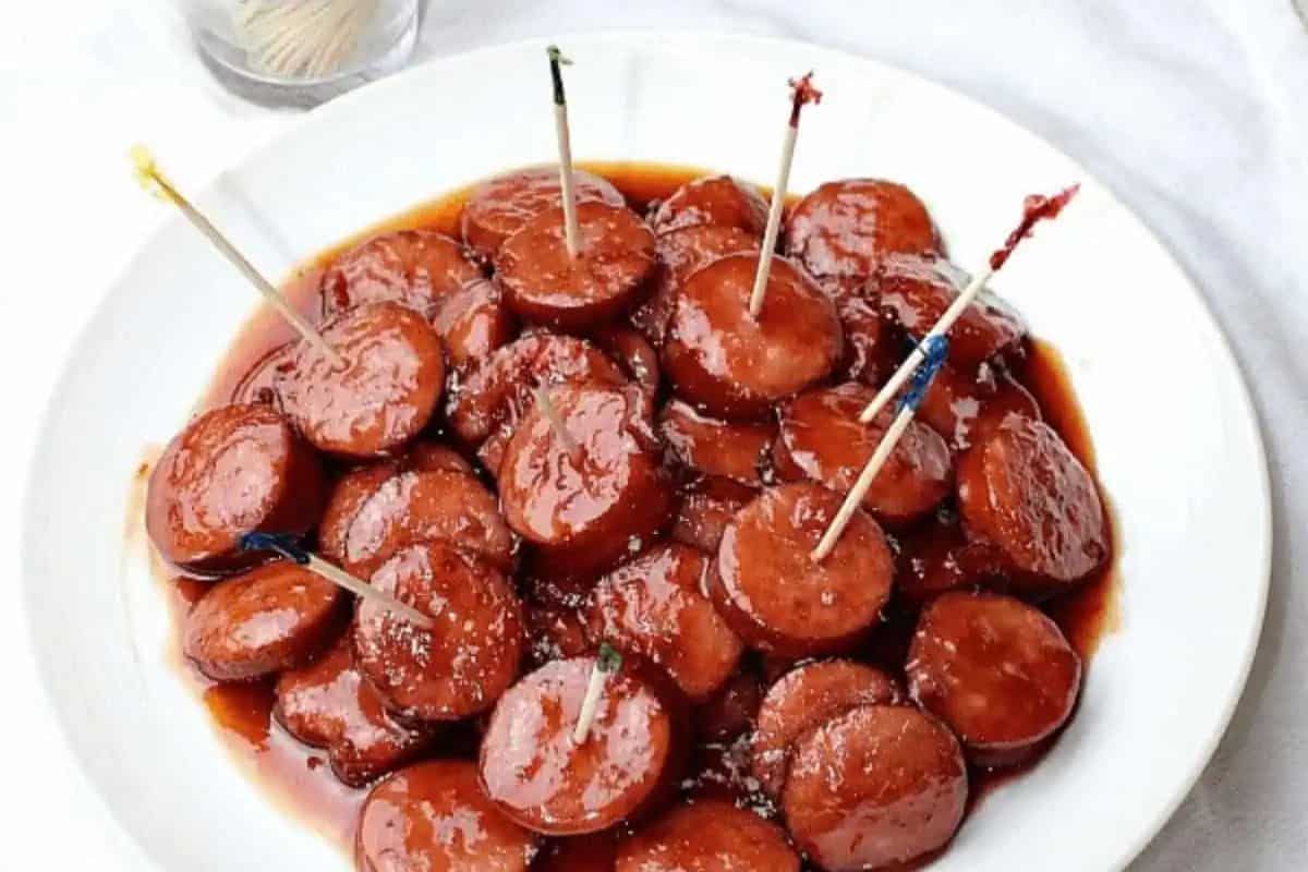 A plate of sliced sausage pieces coated in sauce, each with a toothpick for serving, makes for one of the easiest dinner recipes—perfect for a quick meal or party appetizer. A glass of toothpicks sits in the background.