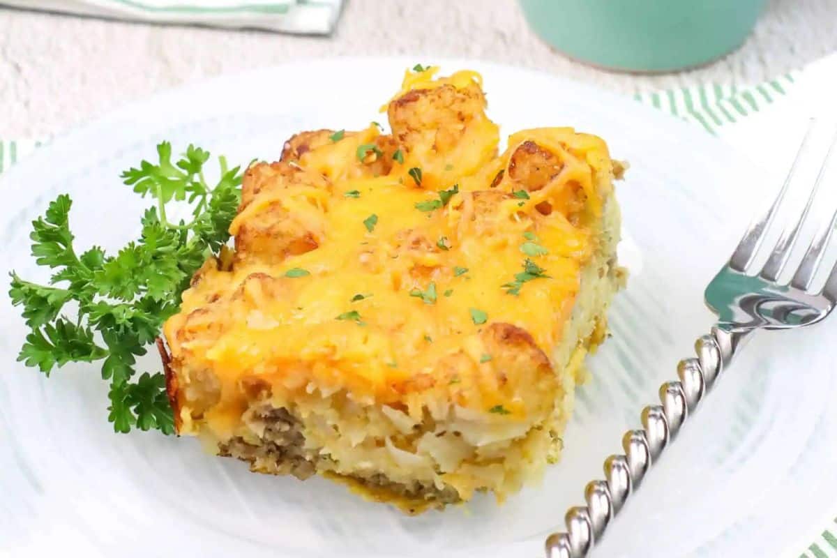 A slice of cheesy casserole topped with tater tots is served on a white plate with a garnish of parsley and a fork beside it—a sweet and savory option perfect for brunch recipes.