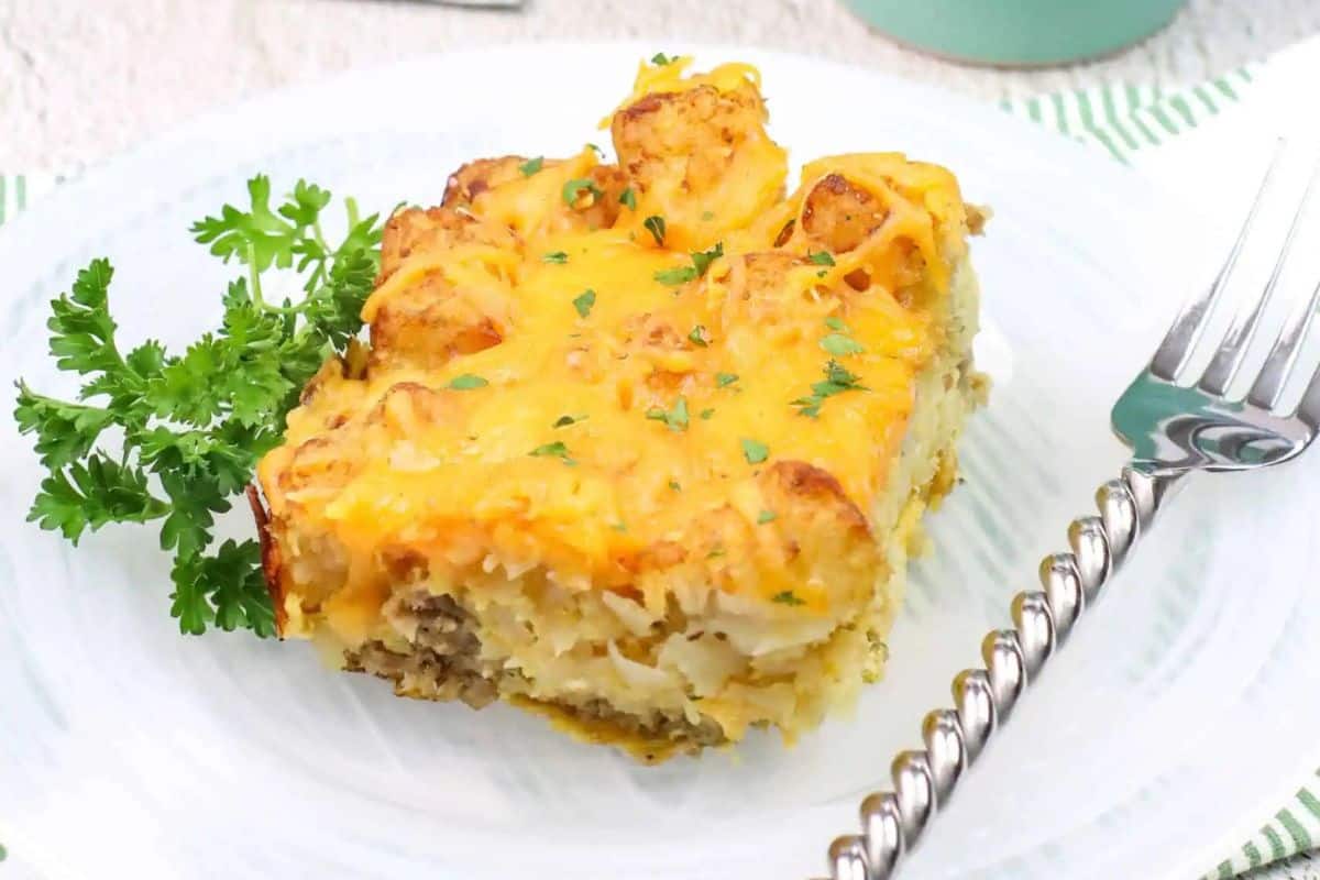 A slice of savory tater tot sausage breakfast casserole with cheese, meat, and a crispy topping rests on a white plate, garnished with parsley for a festive touch. Perfect for brunch or any cheerful St. Patrick's Day gathering, there's even a fork placed beside it, ready to dive in.