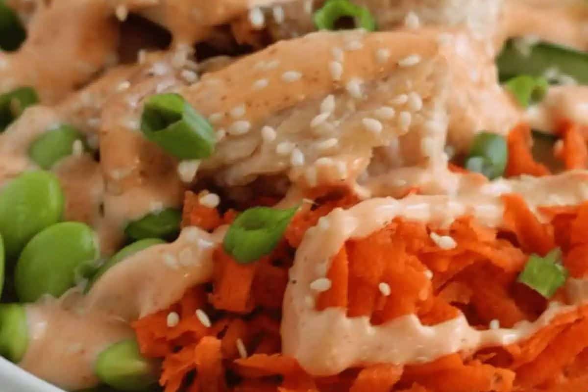 Close-up of a teriyaki salmon bowl with shredded carrots, green edamame, sesame seeds, and a creamy spicy mayo drizzled over irresistible salmon pieces.