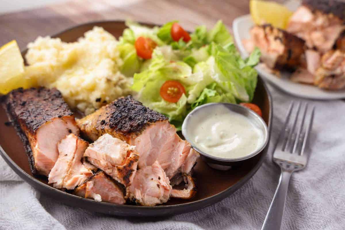 Irresistible grilled salmon with mashed potatoes, a green salad with cherry tomatoes, lemon wedges, and a small container of creamy sauce on a dark plate. A dish from salmon recipes everyone will love!.