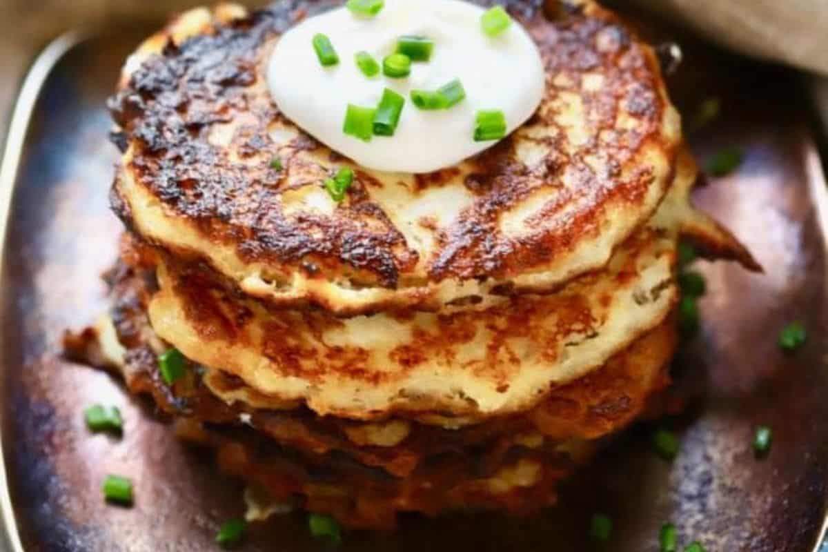 A stack of Irish-inspired potato pancakes topped with sour cream and garnished with chopped chives on a dark plate, delivering a taste of soul-comforting food.