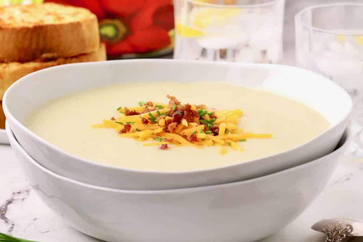 A bowl of creamy, Irish-inspired potato soup topped with shredded cheese, bacon bits, and chives—real comfort food to warm your soul. Glasses of water and slices of toasted bread accompany this heartwarming dish in the background.