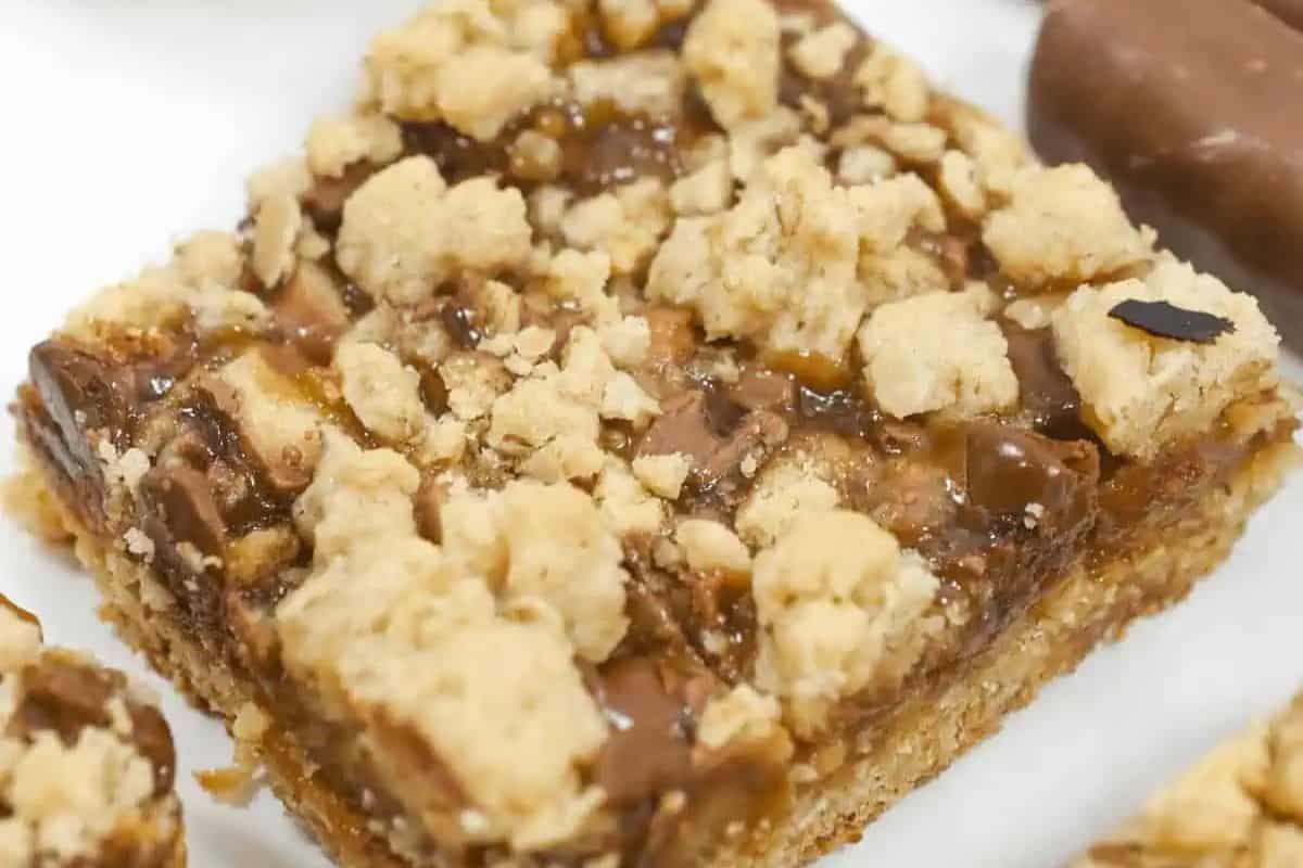 Close-up of a twix oatmeal carmel bar with a caramel and chocolate filling, reminiscent of blondies, topped with a streusel-like crumble on a white background.