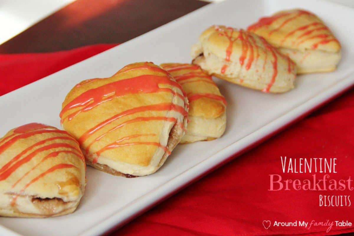 Heart-shaped biscuits with red icing drizzle are arranged on a rectangular white plate, set on a red tablecloth. Perfect for a sweet and savory Valentine's Day breakfast, text on image reads "Valentine Breakfast Biscuits.
