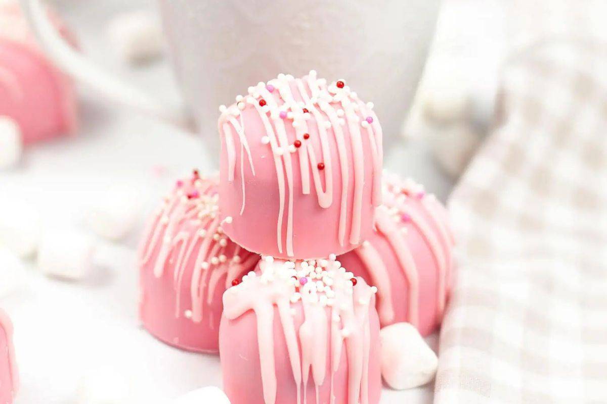 Three pink cocoa bombs with white drizzle and sprinkles are stacked in front of a white cup, with a checkered cloth nearby—perfect for sharing festive recipes on Galentine’s Day.