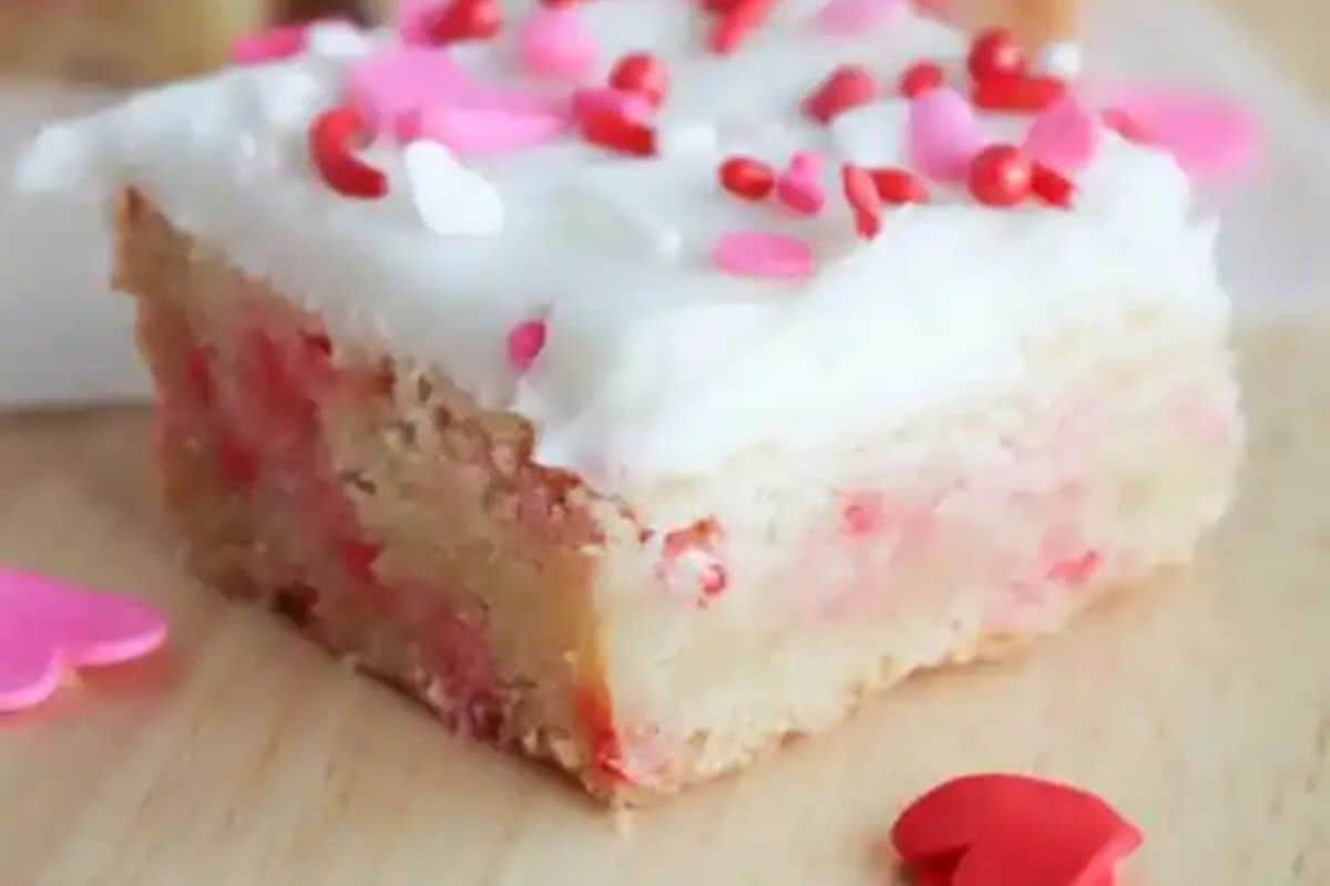 A square slice of a sugar cookie bar with white frosting and pink and red heart-shaped sprinkles on top, perfect for celebrating Galentine’s Day with your bestie, rests on a wooden surface.