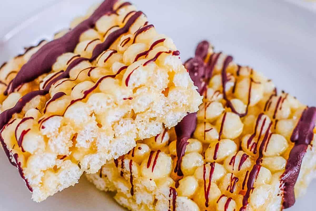 Two stacked vegan rice krispie treats drizzled with purple chocolate on a white plate present an irresistible delight. Perfect for elevating your snack game, these treats showcase the creative potential of Rice Krispie treat recipes.