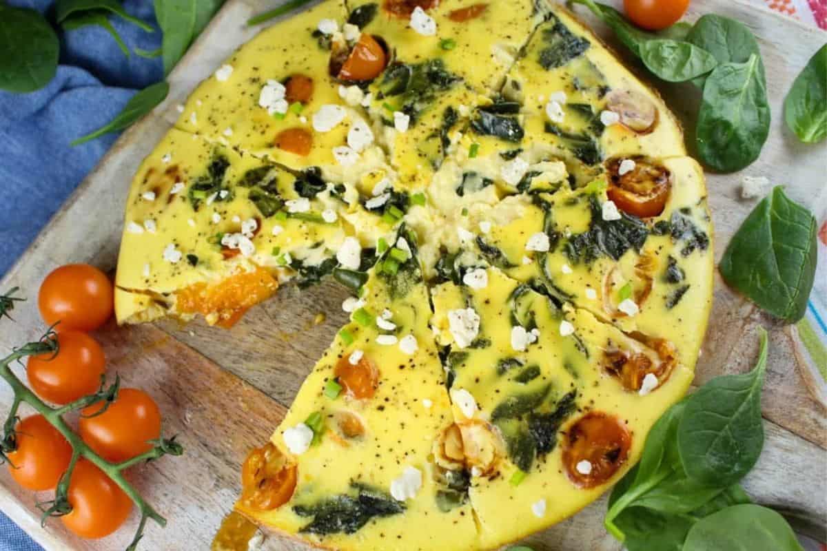 Sliced spinach and tomato frittata garnished with crumbled cheese on a wooden board, surrounded by fresh spinach leaves and cherry tomatoes—perfect for fans of easy Weight Watchers recipes.