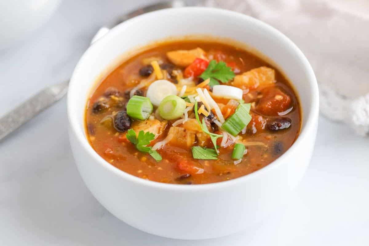 A bowl of chicken tortilla soup topped with sliced green onions, shredded cheese, and cilantro sits on a white surface—a must try dish inspired by classic chili recipes that everyone loves.