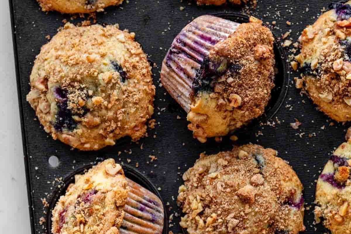 A close-up of blueberry muffins with crumb topping in a black muffin tray, with one muffin slightly tilted in its paper liner—perfect crowd-pleasing desserts and creative bake sale treats.