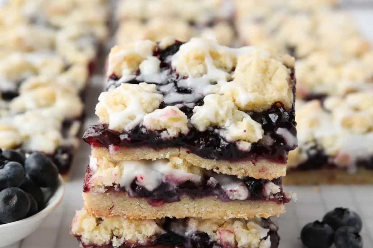 Three stacked blueberry crumb bars with icing drizzle, featuring layers of crumbly topping, blueberry filling, and shortbread base. Fresh blueberries are visible beside these crowd-pleasing desserts—perfect for bake sale treats or creative treats at any gathering.