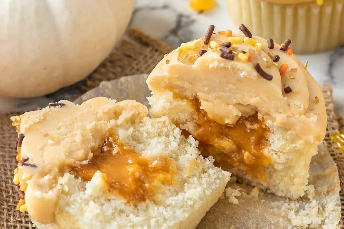 A cupcake cut in half reveals a sweeter caramel filling. The frosting is adorned with brown and yellow sprinkles, while a whole cupcake and a pumpkin sit in the background, perfect for party inspiration or those searching for new cupcake recipes.