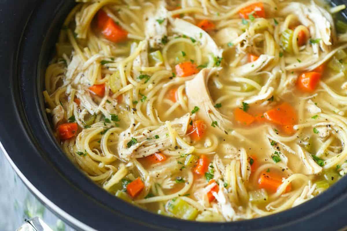 Chicken noodle soup with shredded chicken, carrots, celery, and noodles in broth, garnished with herbs inside a black slow cooker—perfect for your dinner routine or adding to your favorite slow cooker recipes.