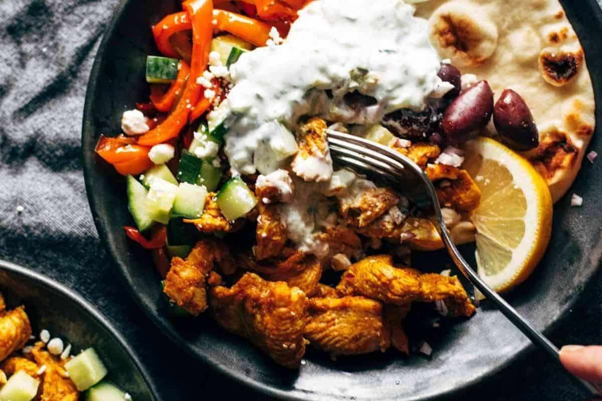 A bowl with delicious grilled chicken, sliced red peppers, cucumber, olives, naan bread, lemon wedge, feta cheese, and a dollop of tzatziki sauce, with a fork picking up food—perfect for easy cleanup after your meal.