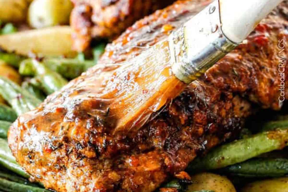 A close-up of a delicious meatloaf being glazed with sauce using a brush, with green beans and potatoes in the background—a perfect example of easy cleanup sheet pan meals.