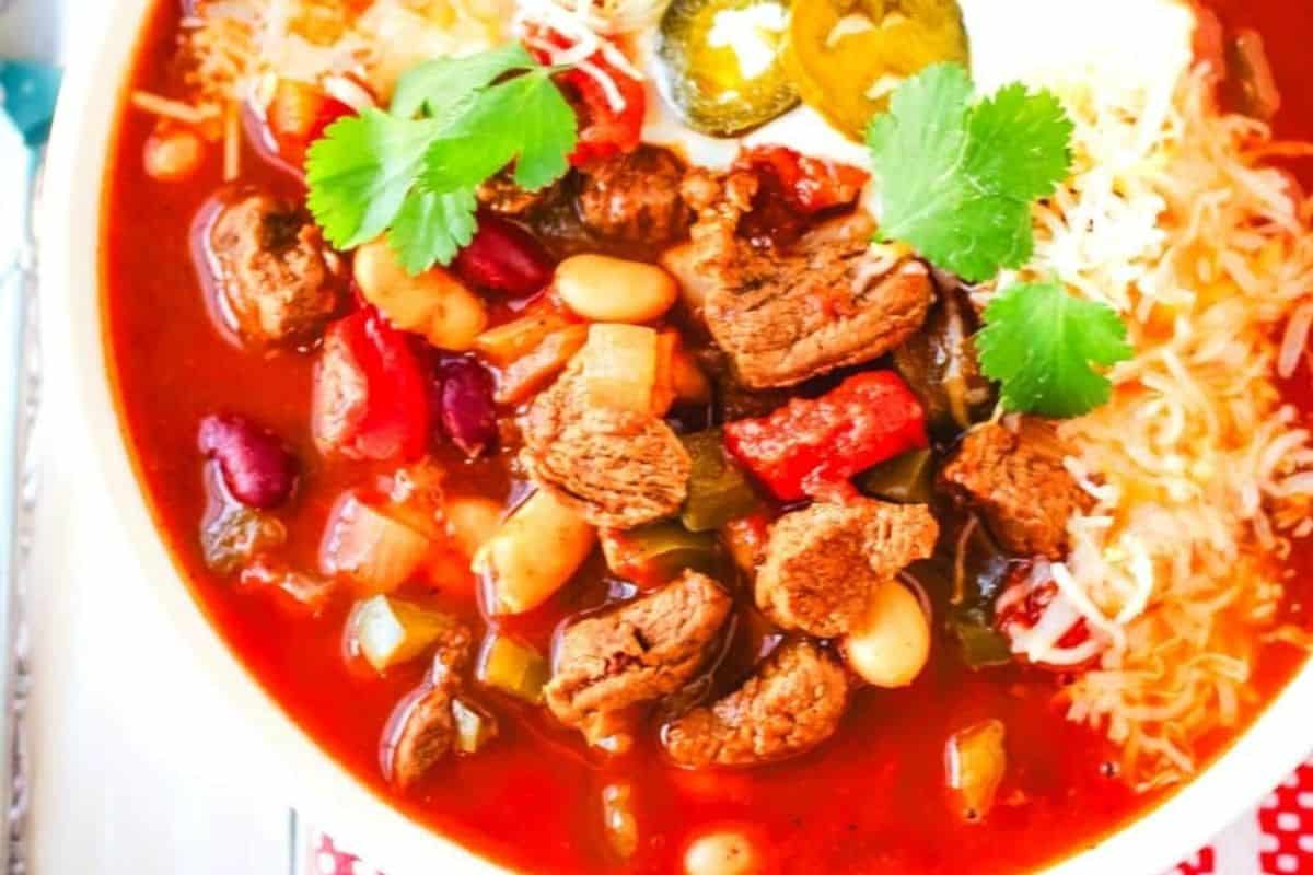 A close-up of a bowl of chili with chunks of beef, beans, tomatoes, shredded cheese, jalapeño slices, sour cream, and fresh cilantro garnish—a must try for fans of classic chili recipes.