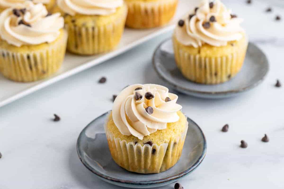 A cupcake with cream frosting and chocolate chips sits on a gray plate, surrounded by scattered chocolate chips, perfect for your next party. More cupcakes await in the background, tempting you to explore sweeter cupcake recipes.