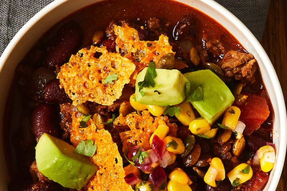 A bowl of chili topped with avocado chunks, corn, red onions, herbs, and crispy cheese pieces—this is one of those chili recipes everyone loves and a must try for comfort food fans.