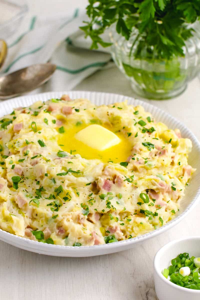 A bowl of mashed potatoes with chopped ham and herbs, topped with melted butter, sits on a table beside a small dish of green onions and a parsley bouquet. Perfect for St. Patrick's Day celebrations, this dish brings an Irish-inspired dinner to life.