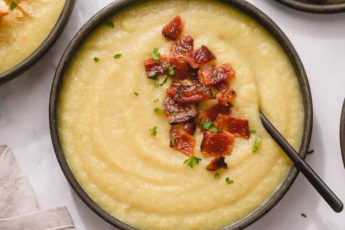 A bowl of creamy yellow soup—one of those comforting soups—topped with crispy bacon bits and chopped herbs, with a black spoon resting inside.
