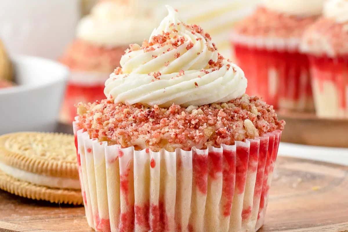 Close-up of a party-ready cupcake with white frosting and red crumbs on top. Nestled in a paper liner, it invites you to explore sweeter delights, with blurred cookies and more cupcakes forming the tempting backdrop.