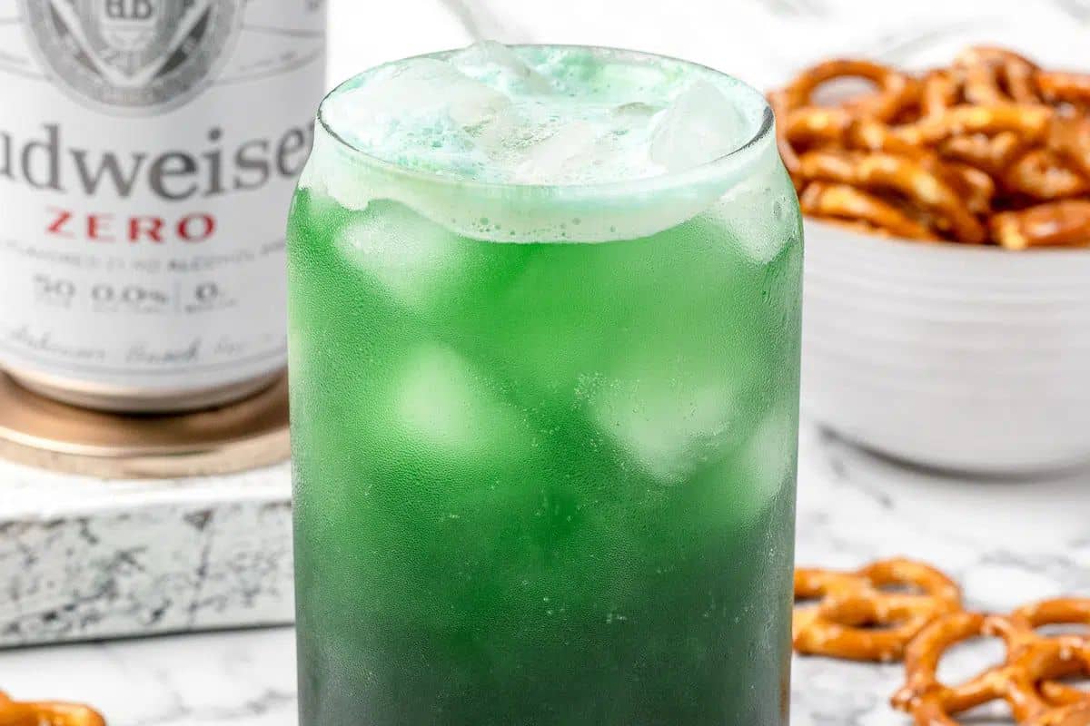 A glass filled with a green iced beverage next to a can of Budweiser Zero and a bowl of pretzels embodies the Irish spirit. A St. Patrick’s Day drink that is a must-try for anyone looking to celebrate in style!.