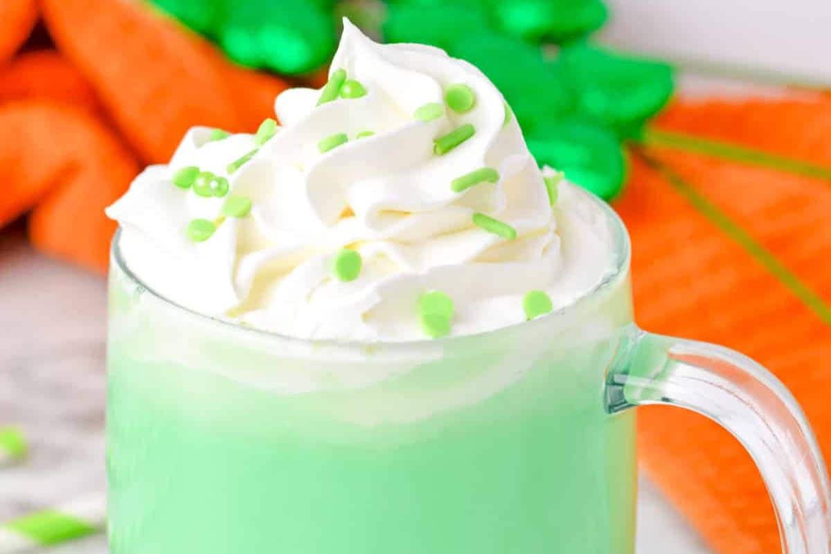 A glass mug filled with a green liquid, topped with whipped cream and green sprinkles, embodies the Irish Spirit. Set against a soft focus background of orange cloth and shiny green decorations, this must-try drink adds a festive touch to St. Patrick's Day celebrations.