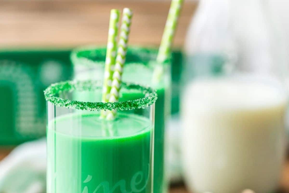 Two glasses of green milk with sugar rims and striped straws evoke the Irish spirit, perfect for a festive St. Patrick’s Day celebration, next to a blurred glass bottle.