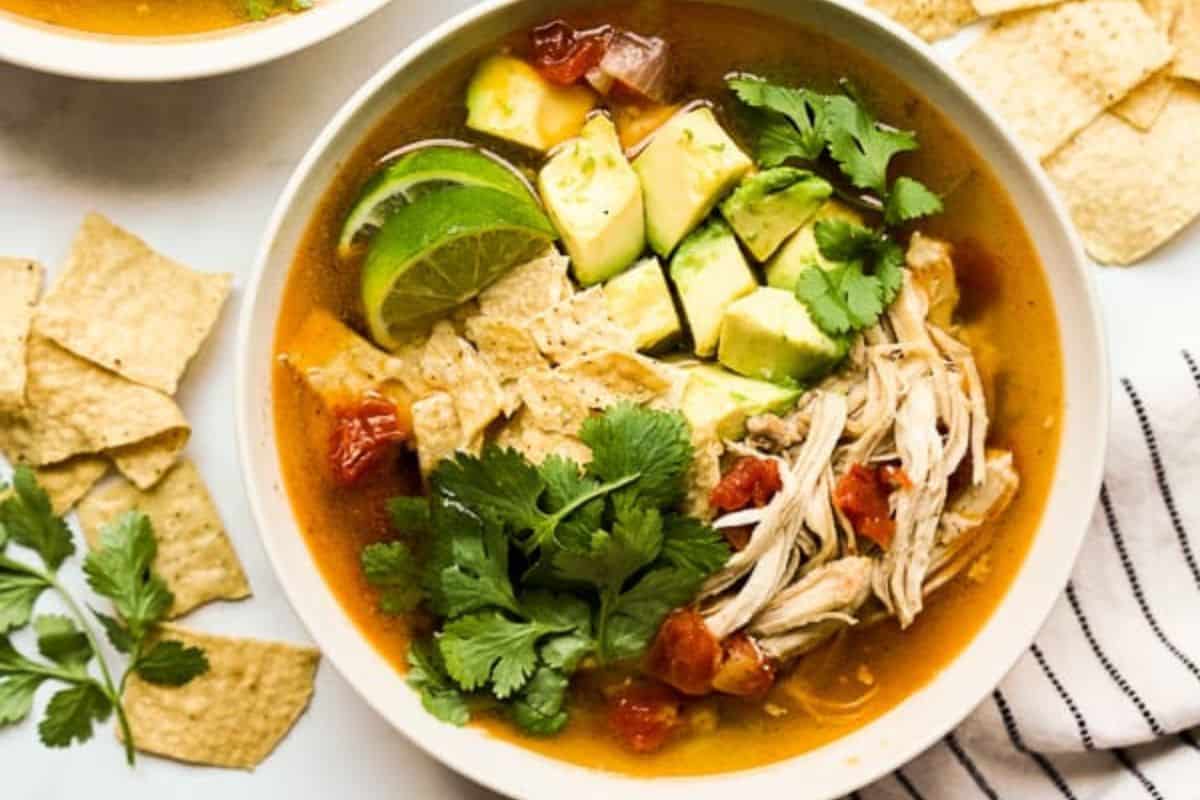 A bowl of chicken tortilla soup topped with avocado cubes, lime slices, cilantro, and shredded chicken, with tortilla chips on the side—one of the best comforting soups you can make in just 30 minutes.