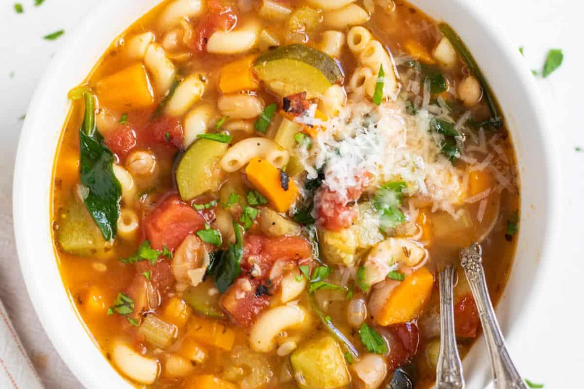 A bowl of vegetable soup with macaroni, diced tomatoes, carrots, zucchini, spinach, white beans, and grated cheese is the perfect example of comforting soups—ready in just 30 minutes and served with two spoons for sharing.