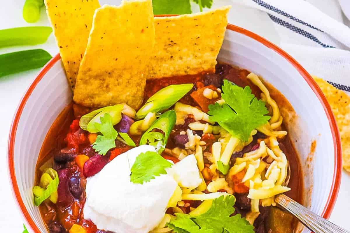 A bowl of popular chili topped with shredded cheese, sour cream, cilantro, and green onions, served with two tortilla chips on the side.