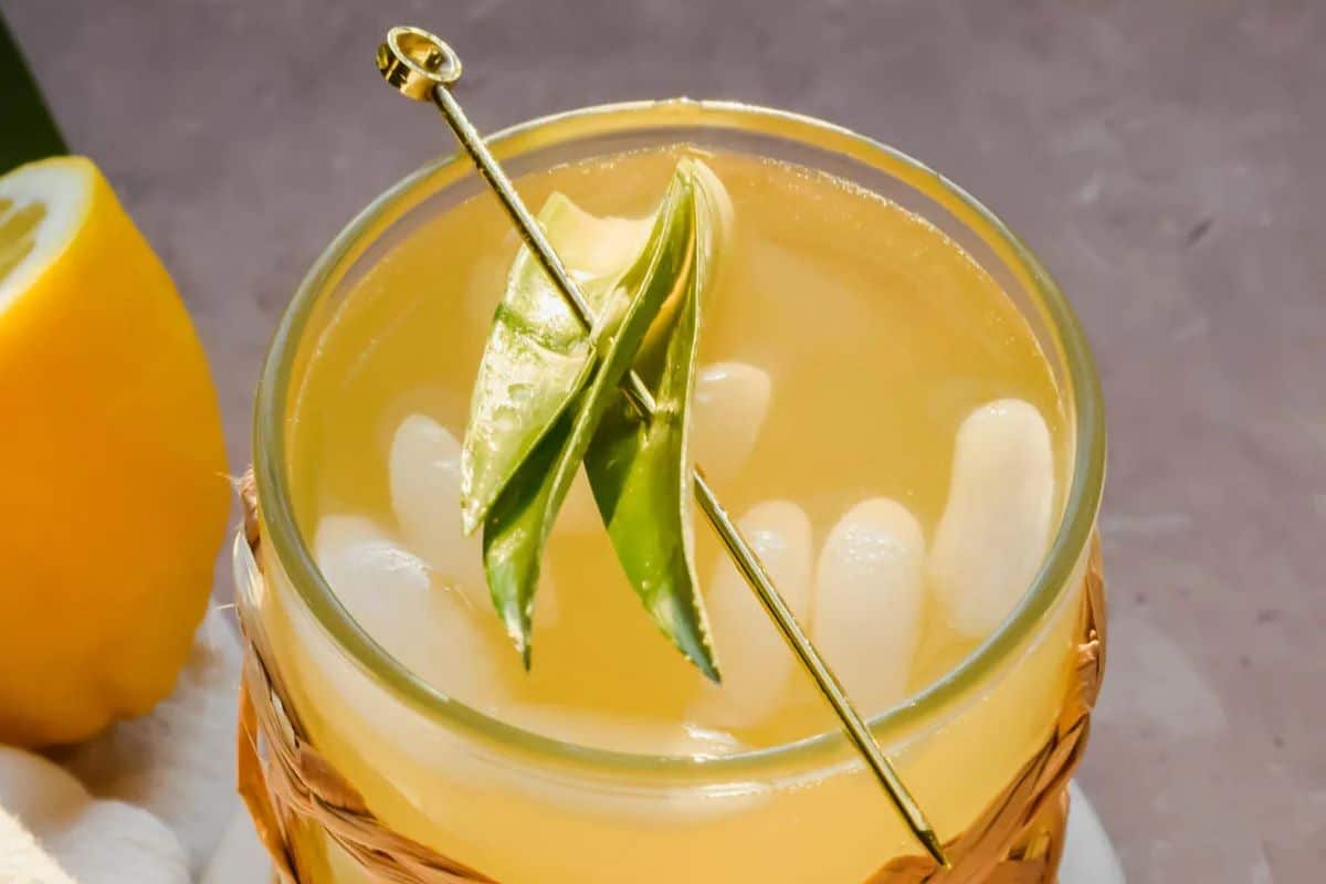 A glass of iced yellow drink, embodying the Irish spirit, is garnished with green leaves on a skewer, placed on a textured surface beside a lemon wedge.