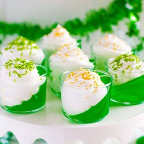 Seven glasses of green gelatin topped with white whipped cream and colored sprinkles are displayed on a white cake stand, embodying the Irish spirit. With green decorations in the background, these are a must-try treat for St. Patrick's Day celebrations.