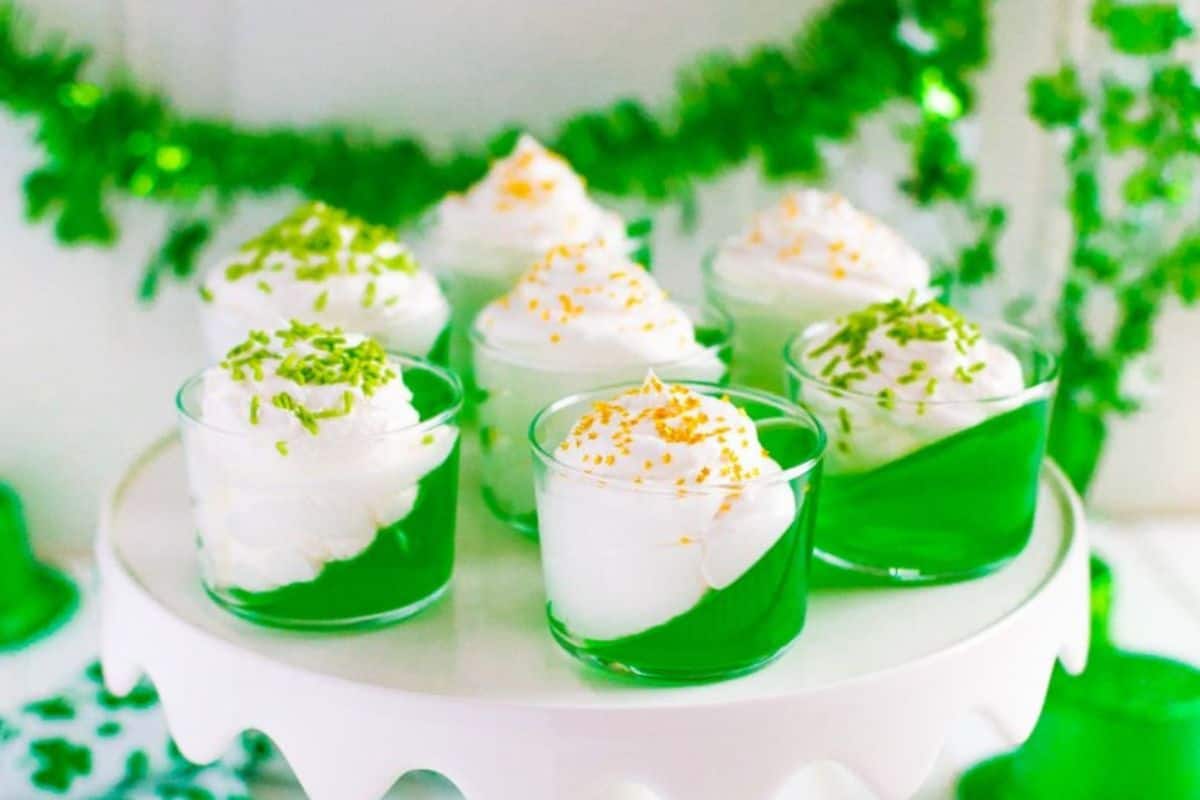 Seven glasses of green gelatin topped with white whipped cream and colored sprinkles are displayed on a white cake stand, embodying the Irish spirit. With green decorations in the background, these are a must-try treat for St. Patrick's Day celebrations.