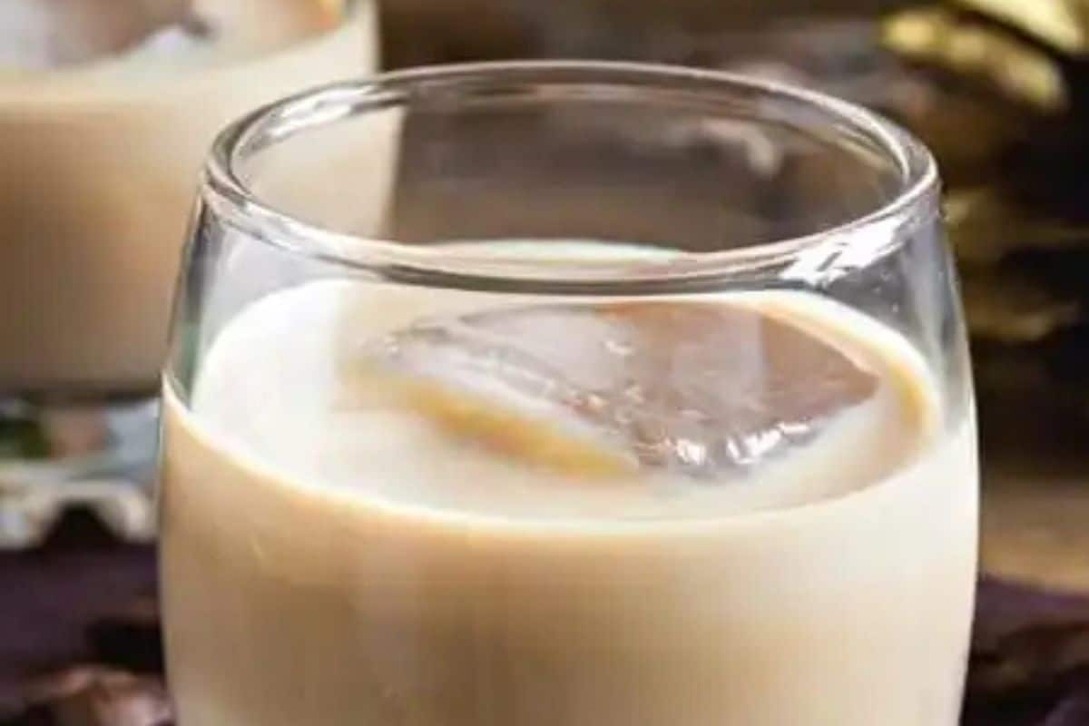 A glass filled with creamy beige liquid containing a large ice cube sits on the table, a perfect Irish spirit to savor during St. Patrick's Day festivities.