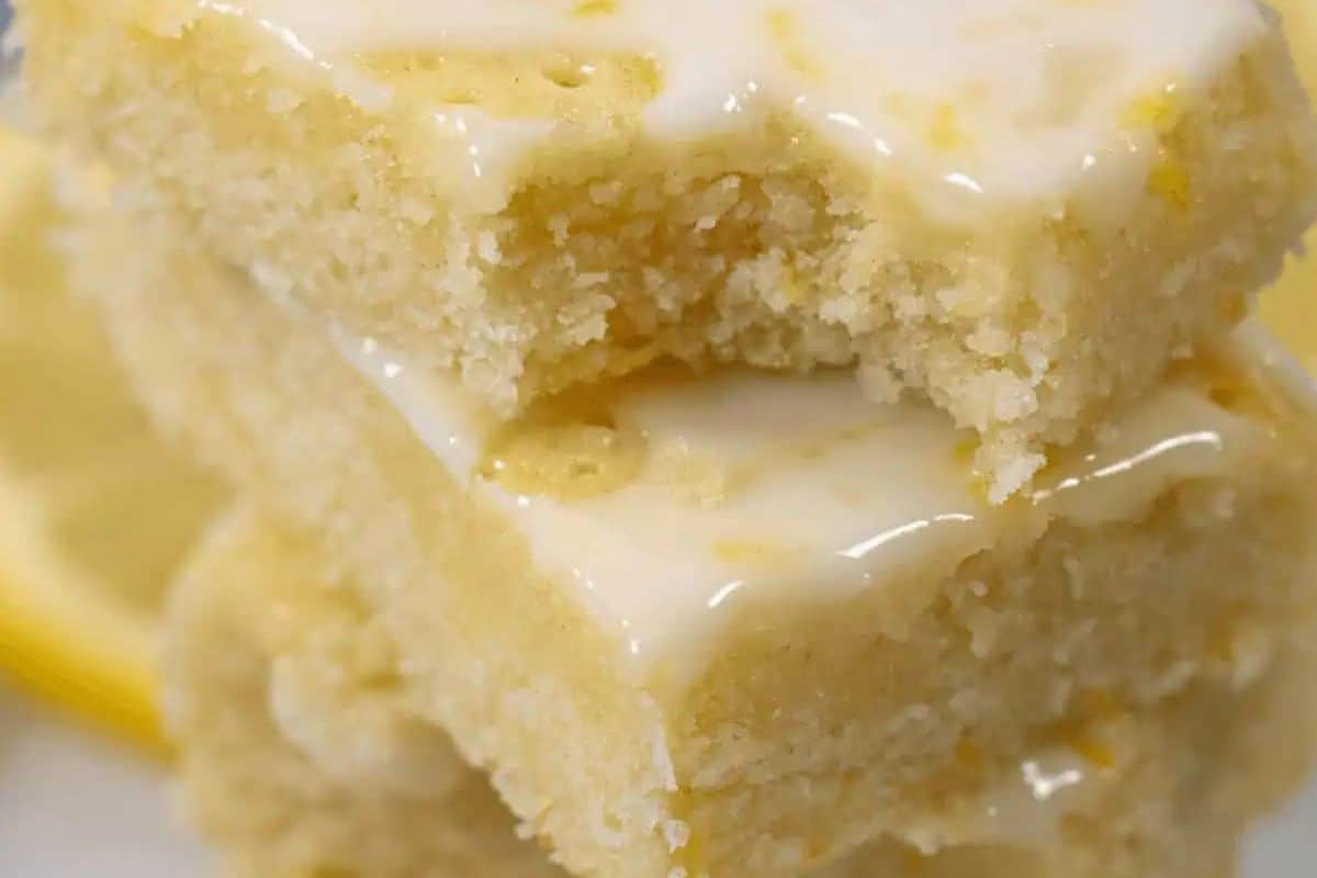 Two stacked lemon bars with a light glaze on top, one with a bite taken out, reveal a moist, dense texture—perfect as crowd-pleasing desserts or creative bake sale treats.