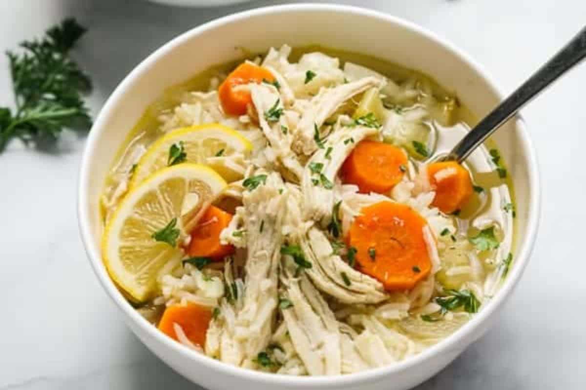 A bowl of chicken soup with shredded chicken, sliced carrots, rice, lemon slices, herbs, and a spoon—one of those comforting soups that's perfect for a cozy meal.