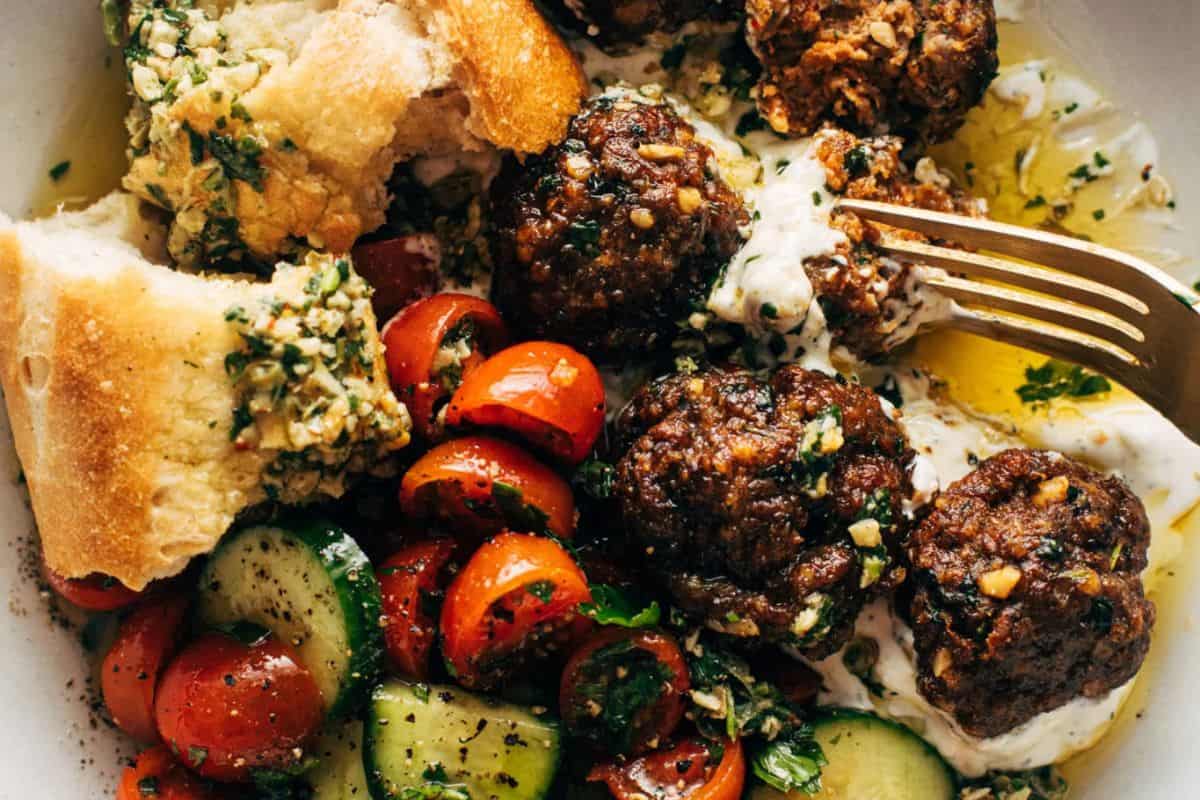 A plate with meatballs on yogurt sauce, sliced cucumber and cherry tomato salad, and pieces of bread for a delicious meal that promises easy cleanup—perfect for fans of Sheet Pan Meals. A fork rests on the side, ready to enjoy.