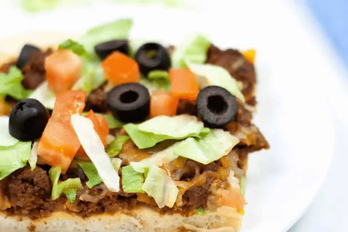 A close-up of a slice of taco pizza topped with ground beef, shredded lettuce, diced tomatoes, sliced black olives, and melted cheese—a Delicious Meal that's perfect for sheet pan meals and easy cleanup.