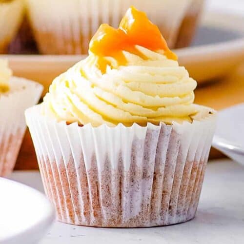 Sweeter cupcake with cream-colored frosting artfully swirled on top, garnished with small orange pieces. Nestled in a white paper liner and placed on a white surface, it's perfect for party gatherings or trying your hand at new cupcake recipes.
