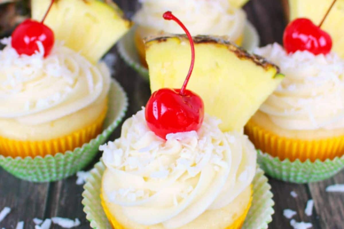 Irresistible cupcakes topped with swirls of frosting, shredded coconut, maraschino cherries, and pineapple wedges, nestled in green paper liners on a wooden surface—a perfect way to make any party sweeter.