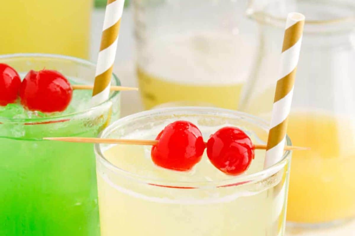 Two glasses of colorful drinks with striped straws evoke an Irish spirit. One drink is green, perfect for St. Patrick’s Day, and the other is yellow, each garnished with two red cherries on skewers. A blurred background features a jug and more beverages.