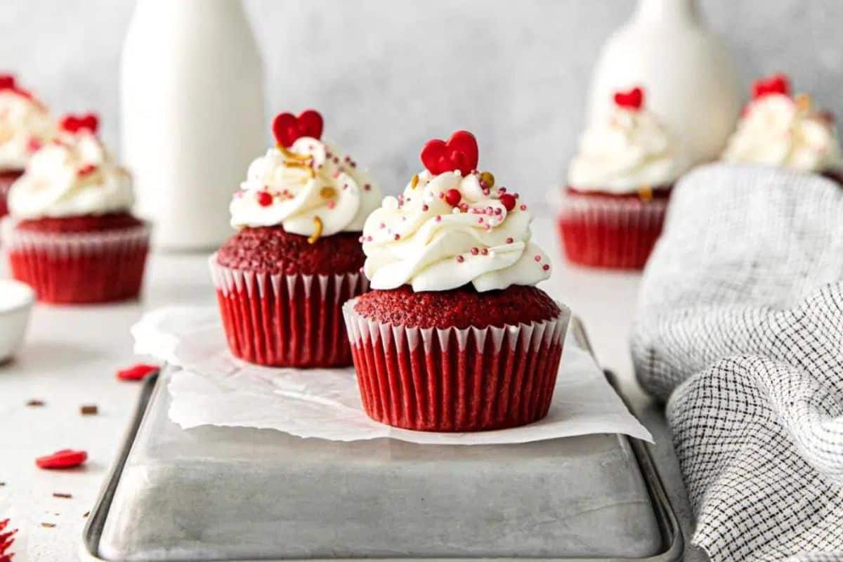 Two red velvet cupcakes with white frosting and red sprinkles sit on a metal tray, tempting any partygoer. More cupcakes and a glass bottle linger in the background, making it feel like the sweetest celebration straight from top-tier cupcake recipes.