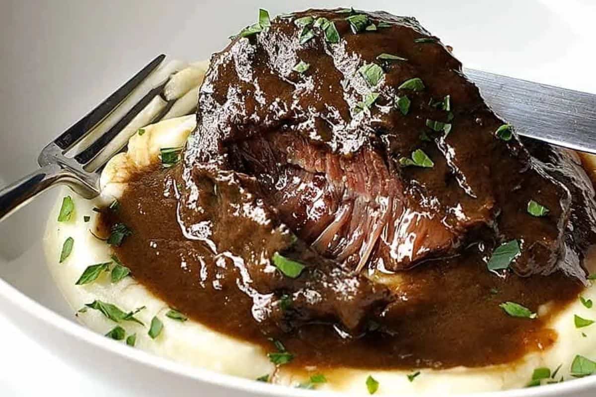 A serving of braised beef in dark gravy sits on mashed potatoes, garnished with chopped parsley, with a fork and knife beside it—a cozy addition to your dinner routine and perfect for fans of hearty dinner recipes.
