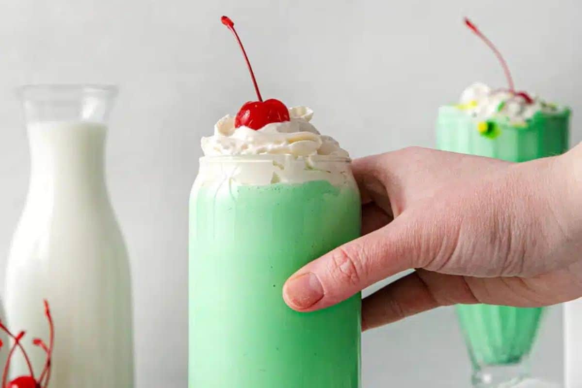A hand holds a light green milkshake topped with whipped cream and a cherry, capturing the Irish spirit of St. Patrick's Day. In the background, a carafe of milk and another similarly adorned drink complete this festive scene.