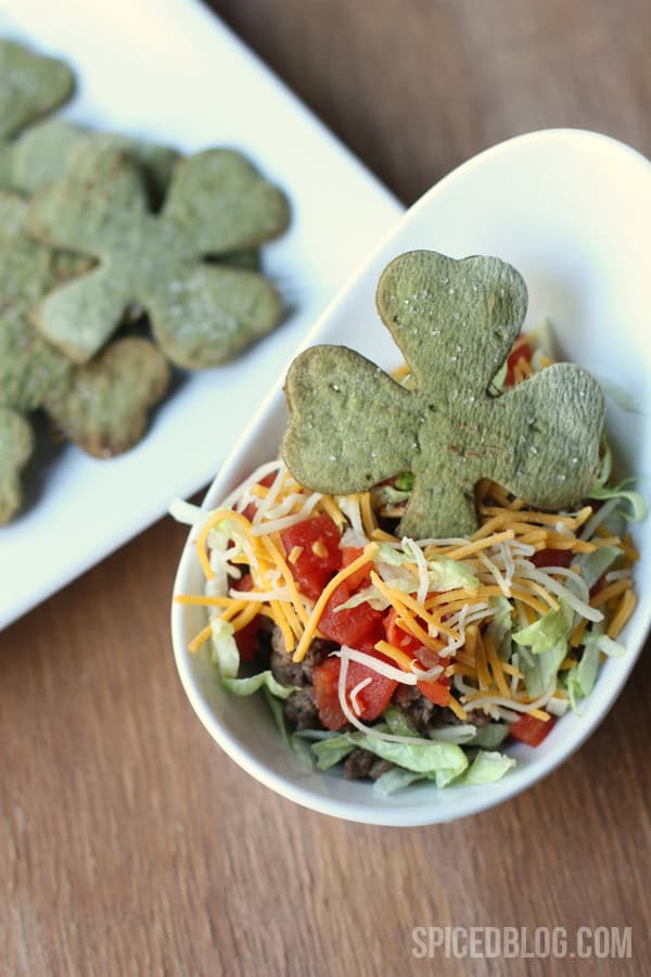 A salad in a white bowl topped with shredded cheese, tomatoes, and lettuce is complemented by a shamrock-shaped green cookie—perfect for St. Patrick's Day. In the background, a plate brims with more festive shamrock cookies, ideal as snacks or embellishments for Irish-inspired dinners.