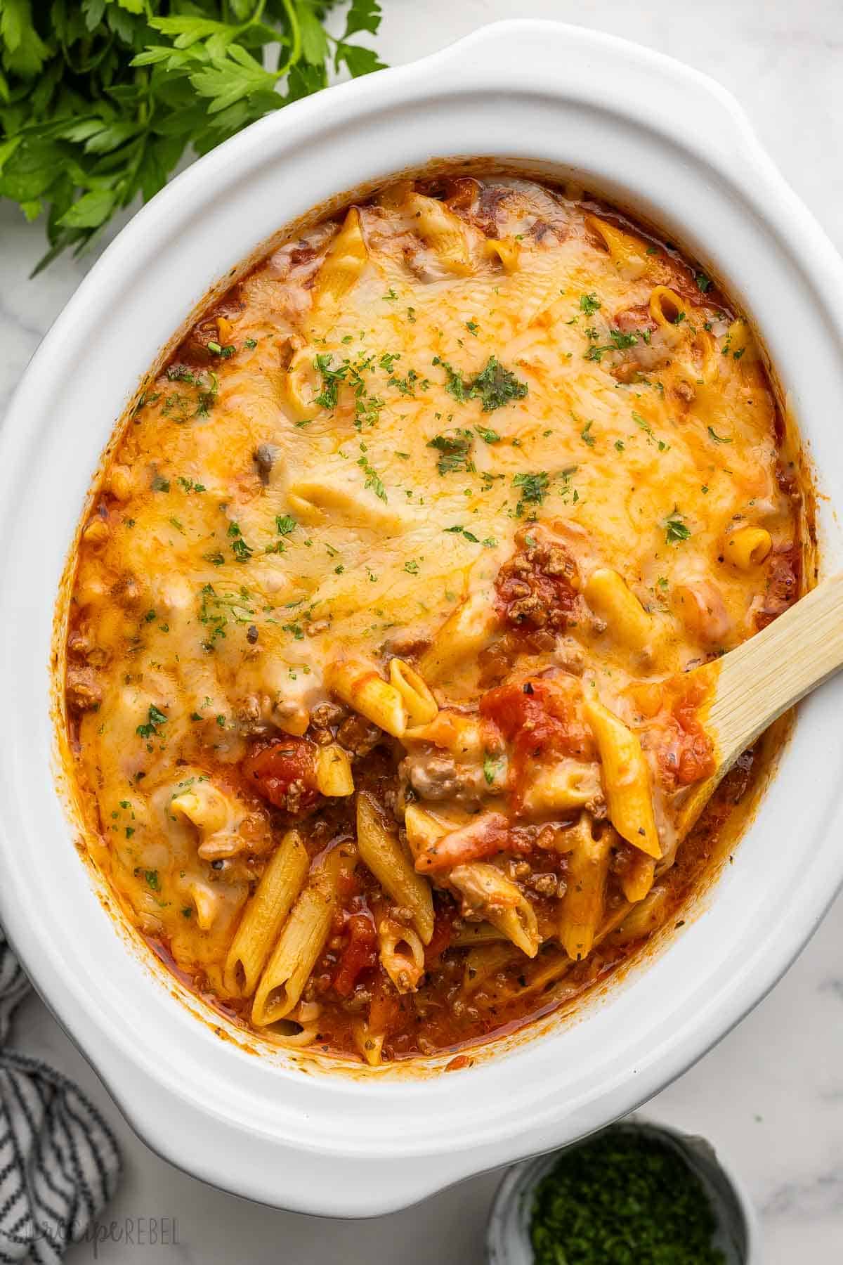 Baked pasta with melted cheese in an oval dish is the perfect potluck recipe. A wooden spoon is partially submerged, inviting you to dive in. Fresh herbs garnish the dish beautifully, making it a delightful addition to any gathering.